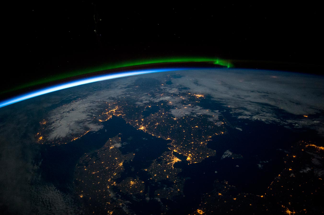 Южная Скандинавия незадолго до полуночи. На фото видно зеленое сияние на севере (верхняя середина изображения), Балтийское море (внизу справа), облака (вверху справа) и снег в Норвегии с подсветкой под полной луной и городские огни на побережье.